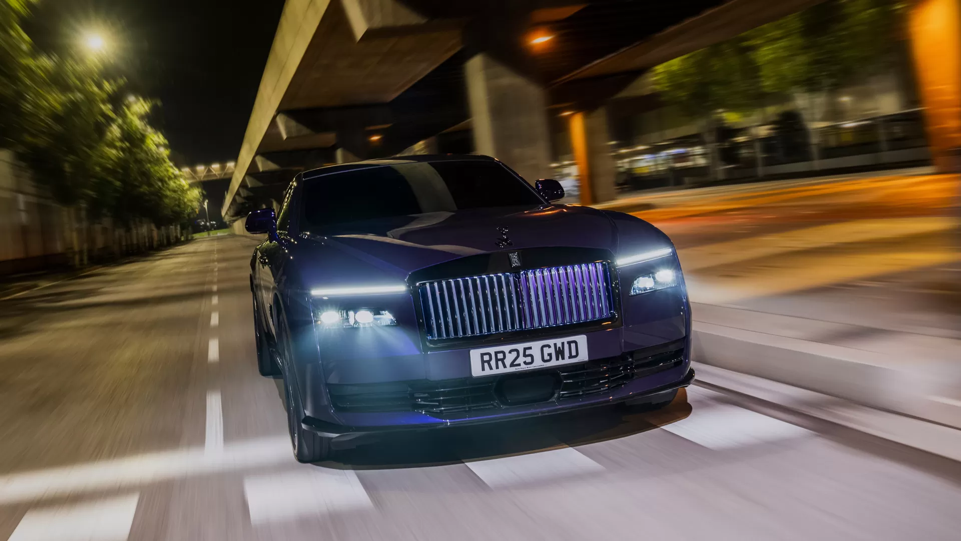 Front View with Iconic Grill: A close-up of the Rolls-Royce Spectre Black Badge showcasing the signature Pantheon grille and Spirit of Ecstasy, surrounded by sleek curves and a polished black finish under dynamic lighting.